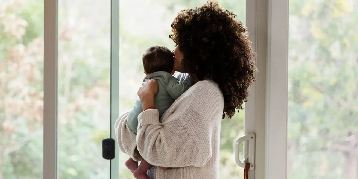 Mom holding baby at 3 months postpartum looking out window during recovery
