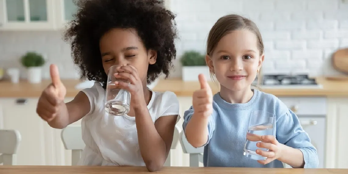Two girls drinking water at home giving thumbs up showing healthy hydration habits that prevent dehydration in kids