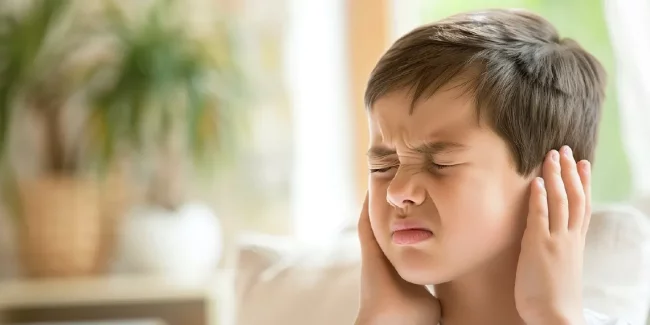 Boy holding ears in pain at home showing symptoms parents need to identify as swimmer's ear or middle ear infection